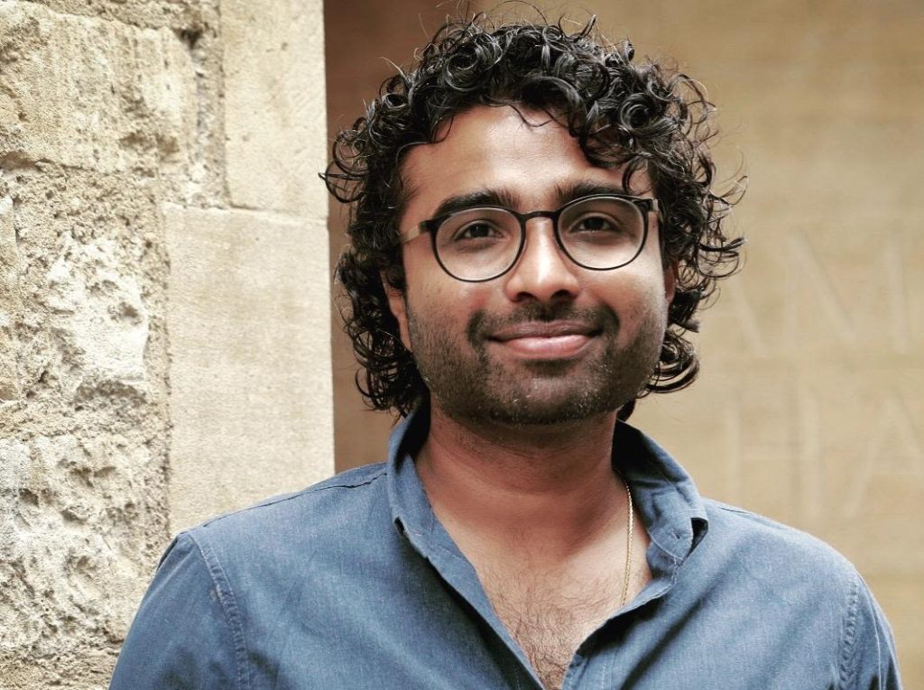 Man with brown skin and dark wavy hair, wearing blue shirt and glasses, stands near wall