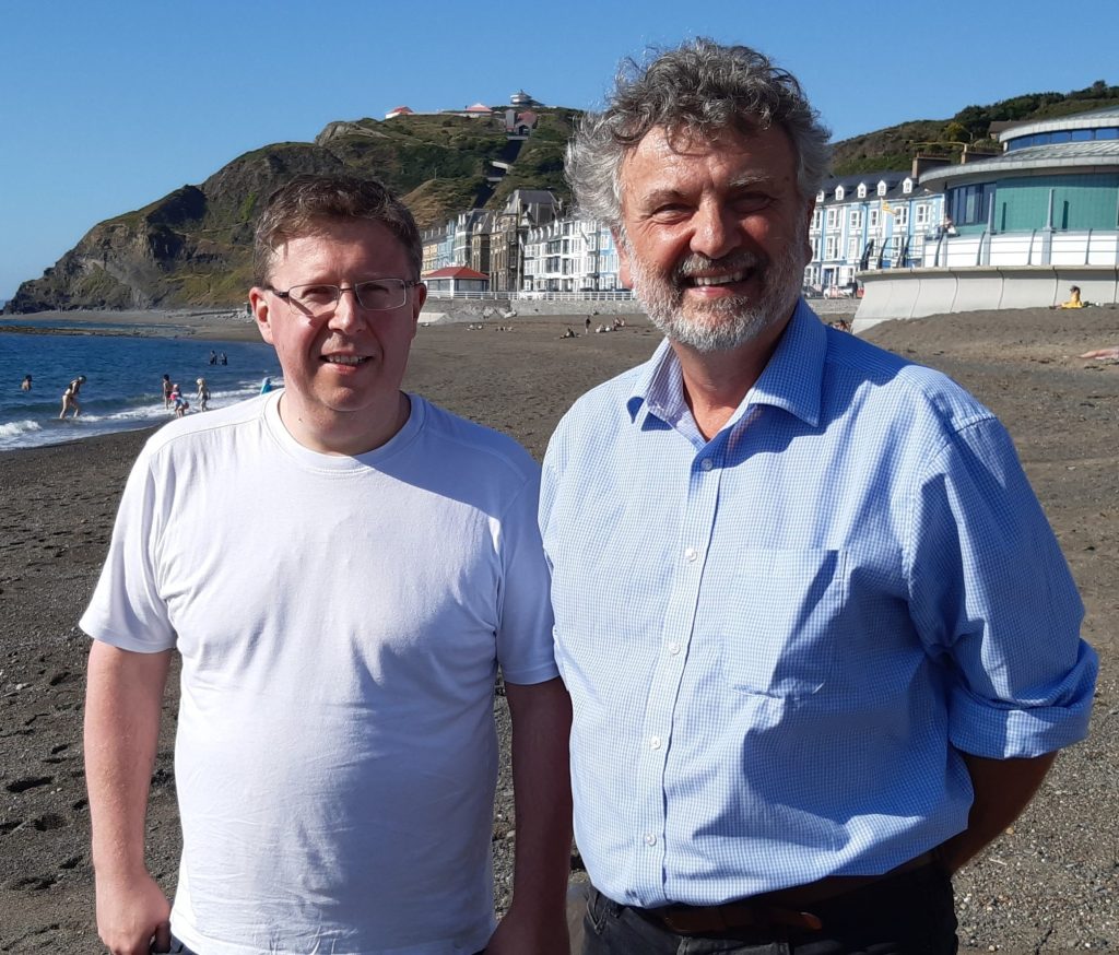 Two men standing on beach smiling, in bay