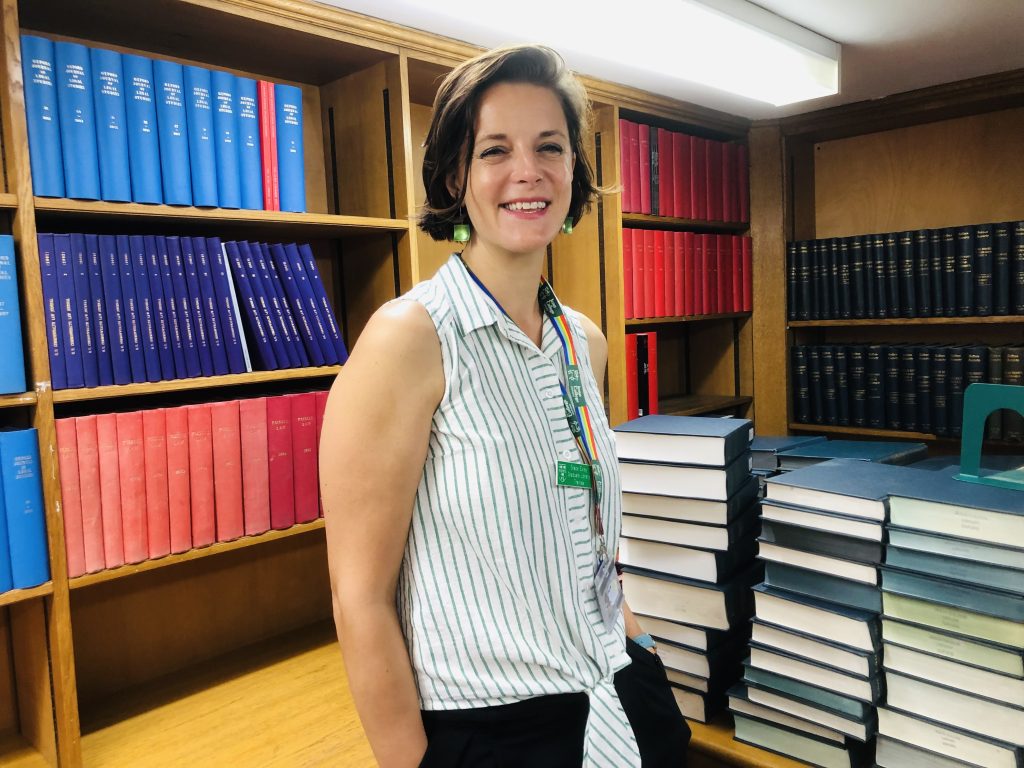 Woman with brown hair and wearing white sleeveless shirt smiles at camera - with books on shelves in background