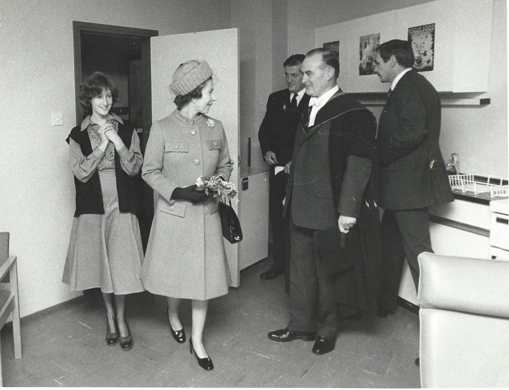 Queen stands in middle of kitchen space with man in university gown on right and smiling female student on left