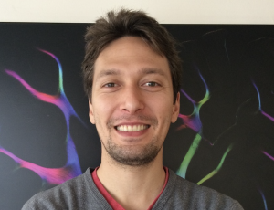 White male with brown hair and slight beard, wearing grey v-neck jumpe