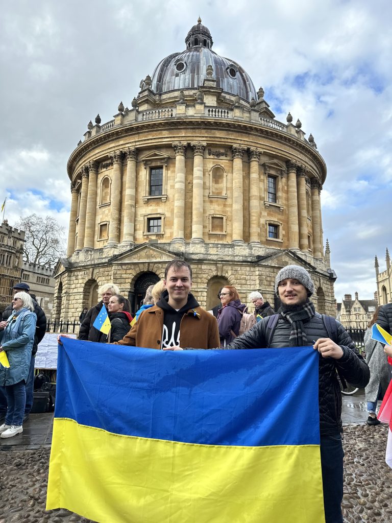 Two men holding a yellow and blue Ukrainian flag, standing in front of the Radcliffe camera. 