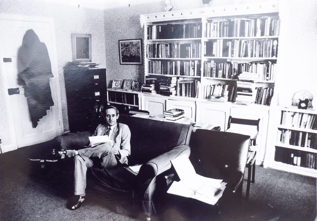 Black and white photo of man in grey suit sitting in a large room with bookshelves in background.