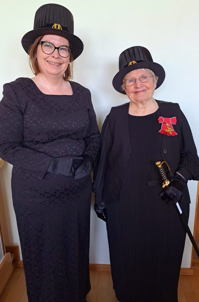 Two women wearing glasses and black hats and dresses smiling at camera. Woman on right - Prof Kathy Sylva - also had red ribbon on dress and is holding a ceremonial sword. 