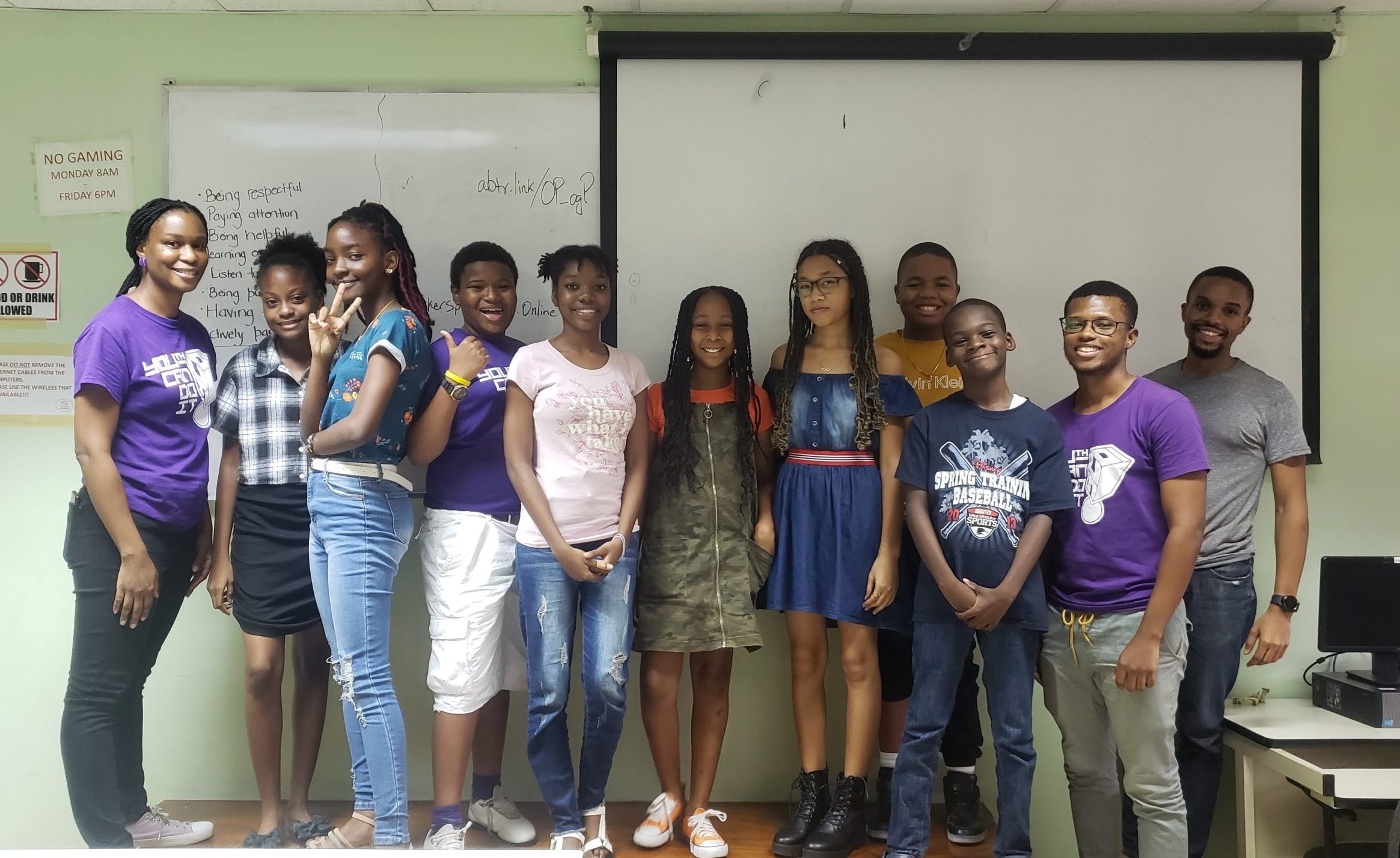 Editoiral: twelve Jamaican young people standing in a row wearing summer clothing, in front of a whiteboard. Smiling to camera.