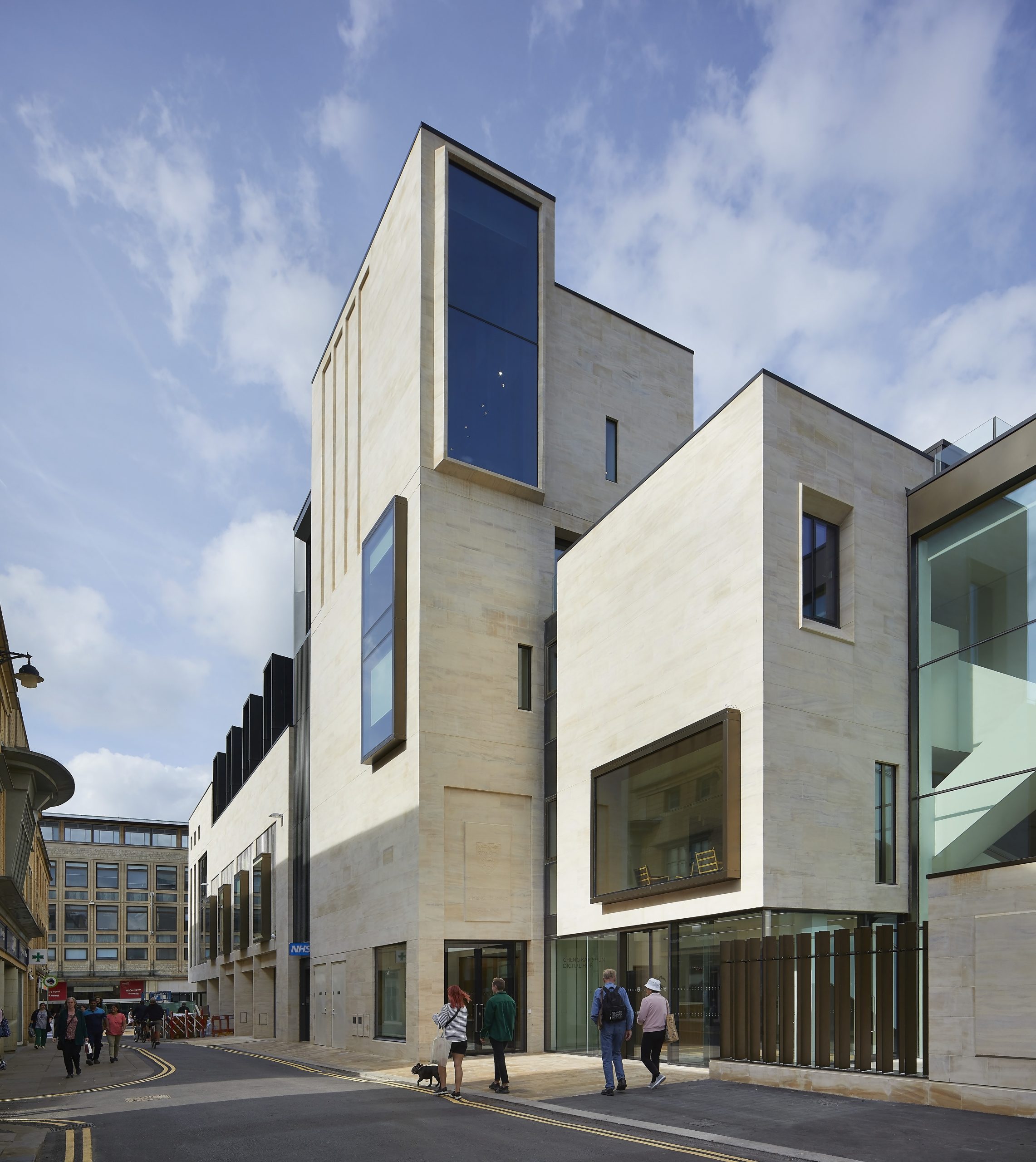 The Cornmarket facade of Jesus College's Cheng Yu Tung Building, designed by MICA Architects. Image shows corner of pale cream stone building with multiple windows and people walking along at ground level Credit: Hufton + Crow