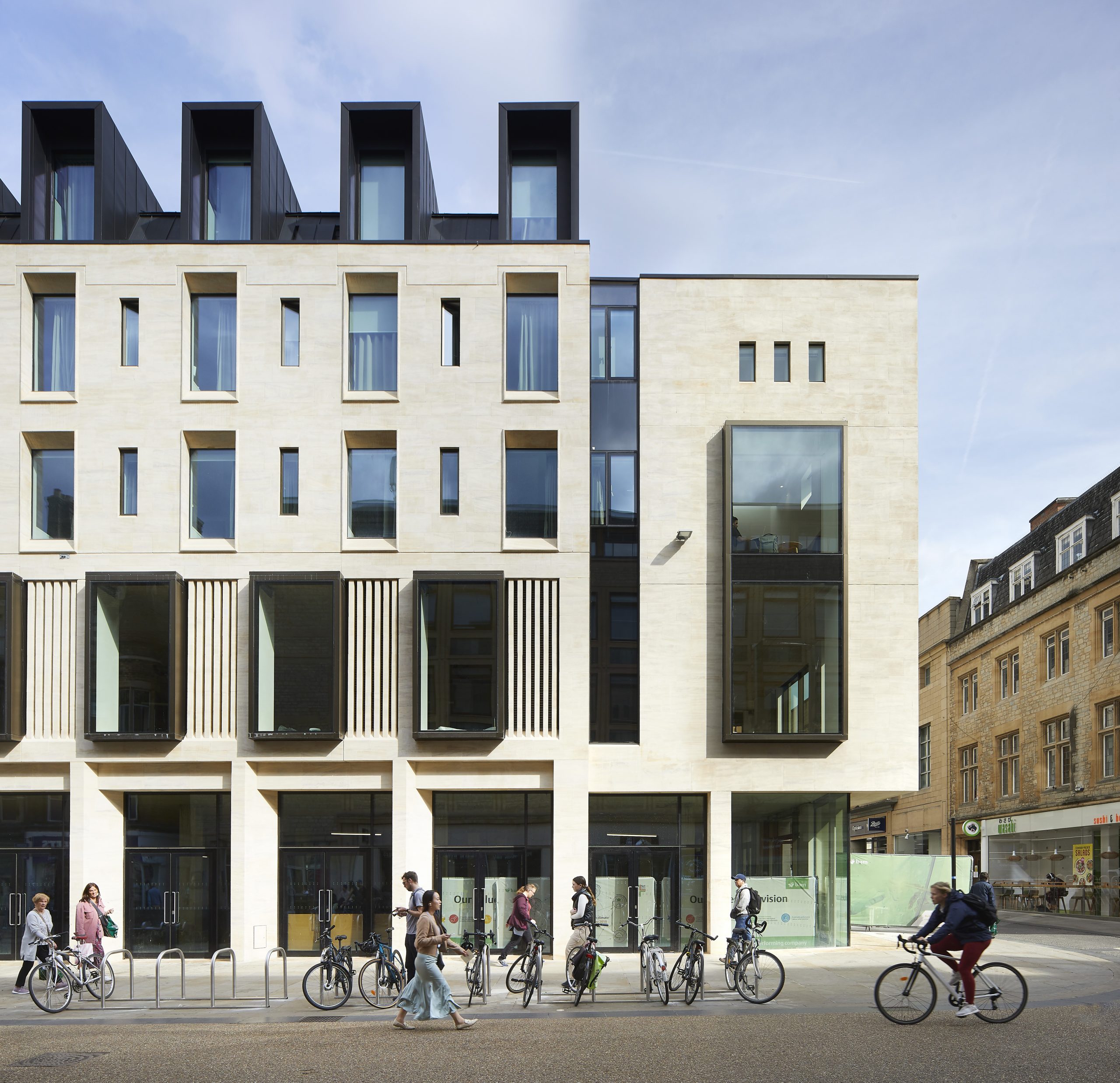 The Cornmarket facade of Jesus College's Cheng Yu Tung Building, designed by MICA Architects. Image shows corner of pale cream stone building with multiple windows and people walking along at ground level Credit: Hufton + Crow