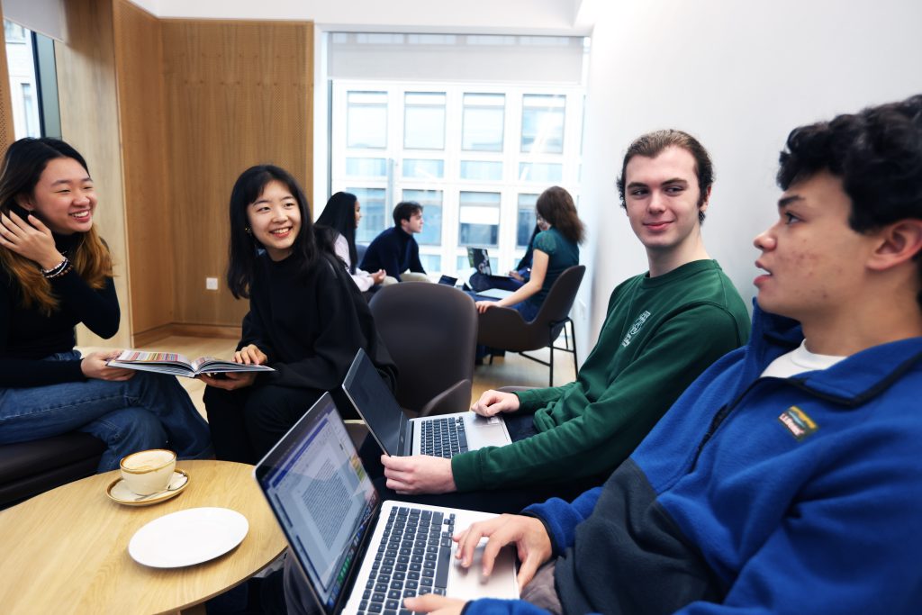 Group of students aged around 19, each wearing different colour tops sit around a wooden low coffee table talking. 