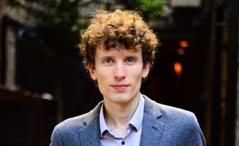 White male with brown curly hair wearing grey suit jacket stands in front of library shelves.