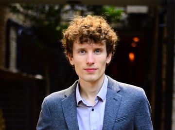 White male with brown curly hair wearing grey suit jacket stands in front of library shelves.