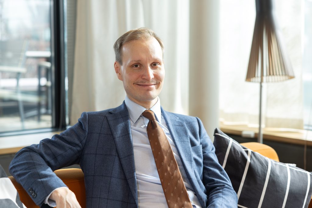 Man in suit and tie with short blonde hair sits on sofa smiling at camera