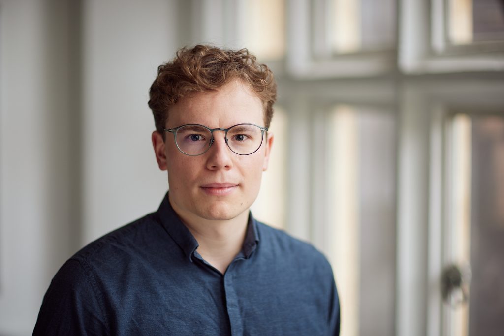 White male wearing glasses with short brown hair and blue shirt stands in front of a window