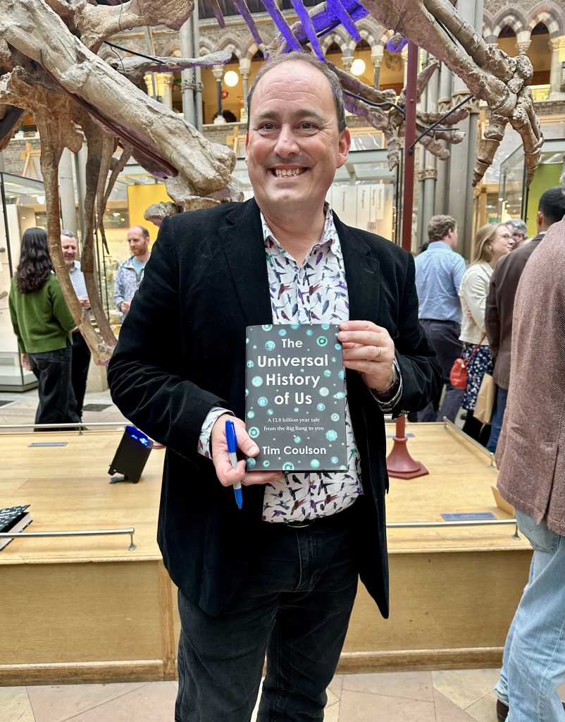 Man in black jacket and patterned shirt smiles at camera holding a book. 