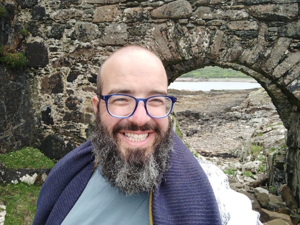 man with grey beard and glasses stands smiling in front of old stone bridge